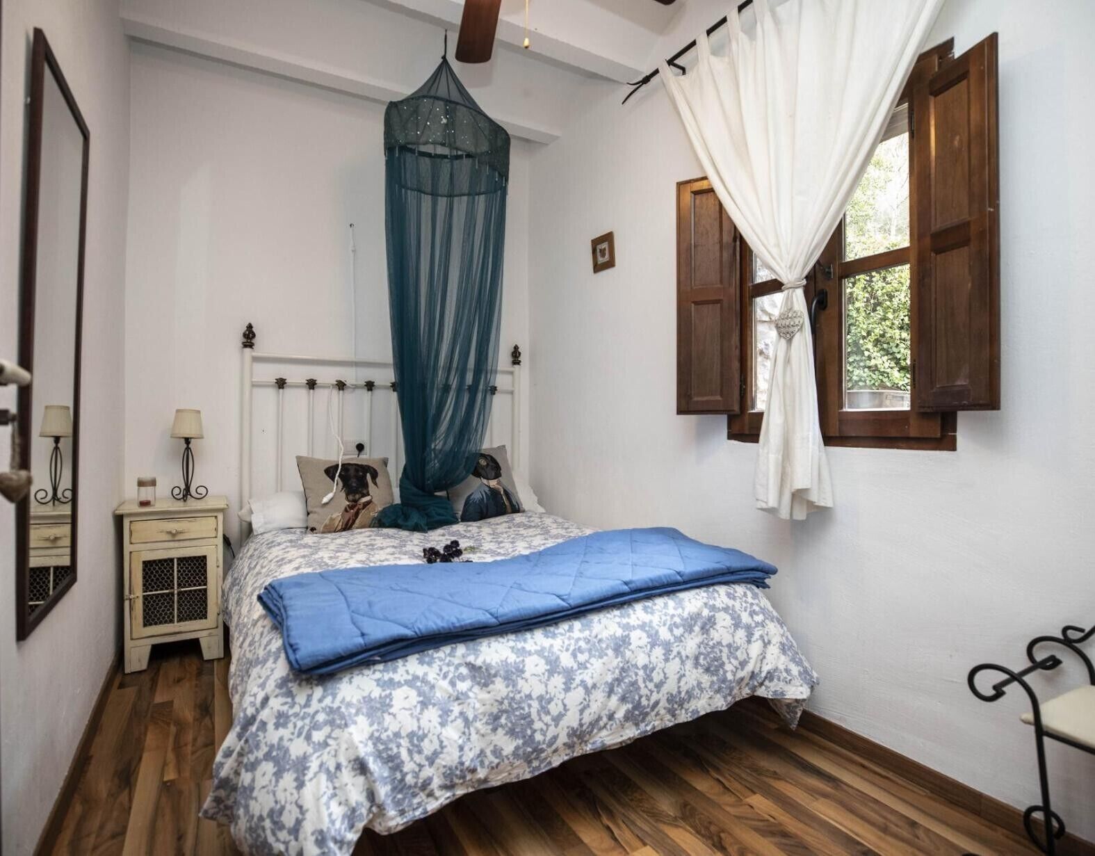 Un dormitorio con cama, mesita de noche, ventana con cortinas y suelo de madera.