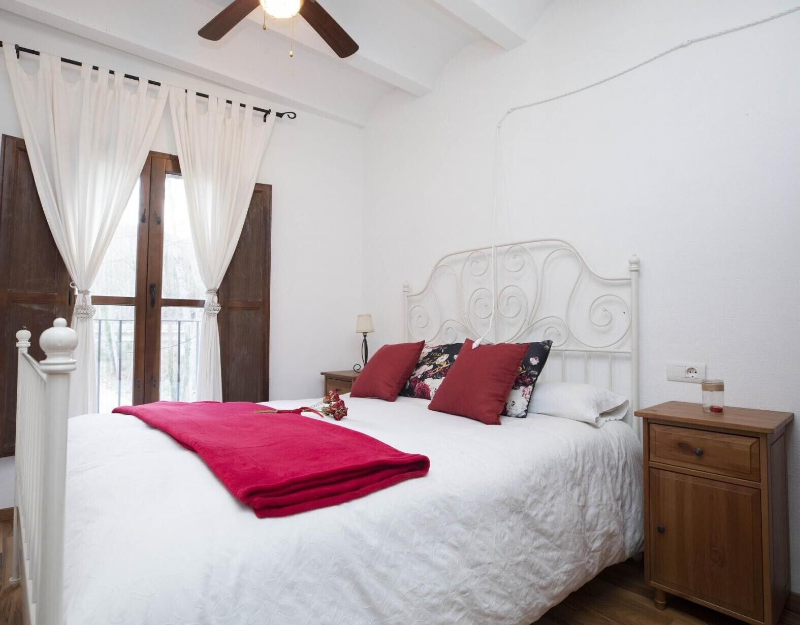 A bedroom with a large bed, white curtains, a wooden nightstand, and a ceiling fan.