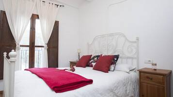 A bedroom with a large bed, white curtains, a wooden nightstand, and a ceiling fan.