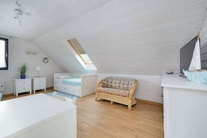 Une pièce avec un plafond en pente, un canapé en osier, un lit blanc, une petite table et une fenêtre donnant sur un beau paysage.