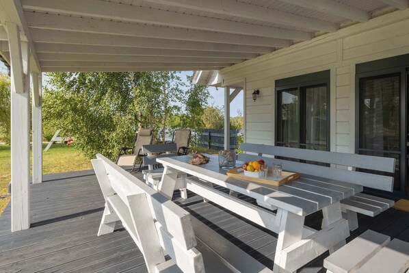 A covered outdoor patio with a wooden table and benches, a small garden area, and a house in the background.