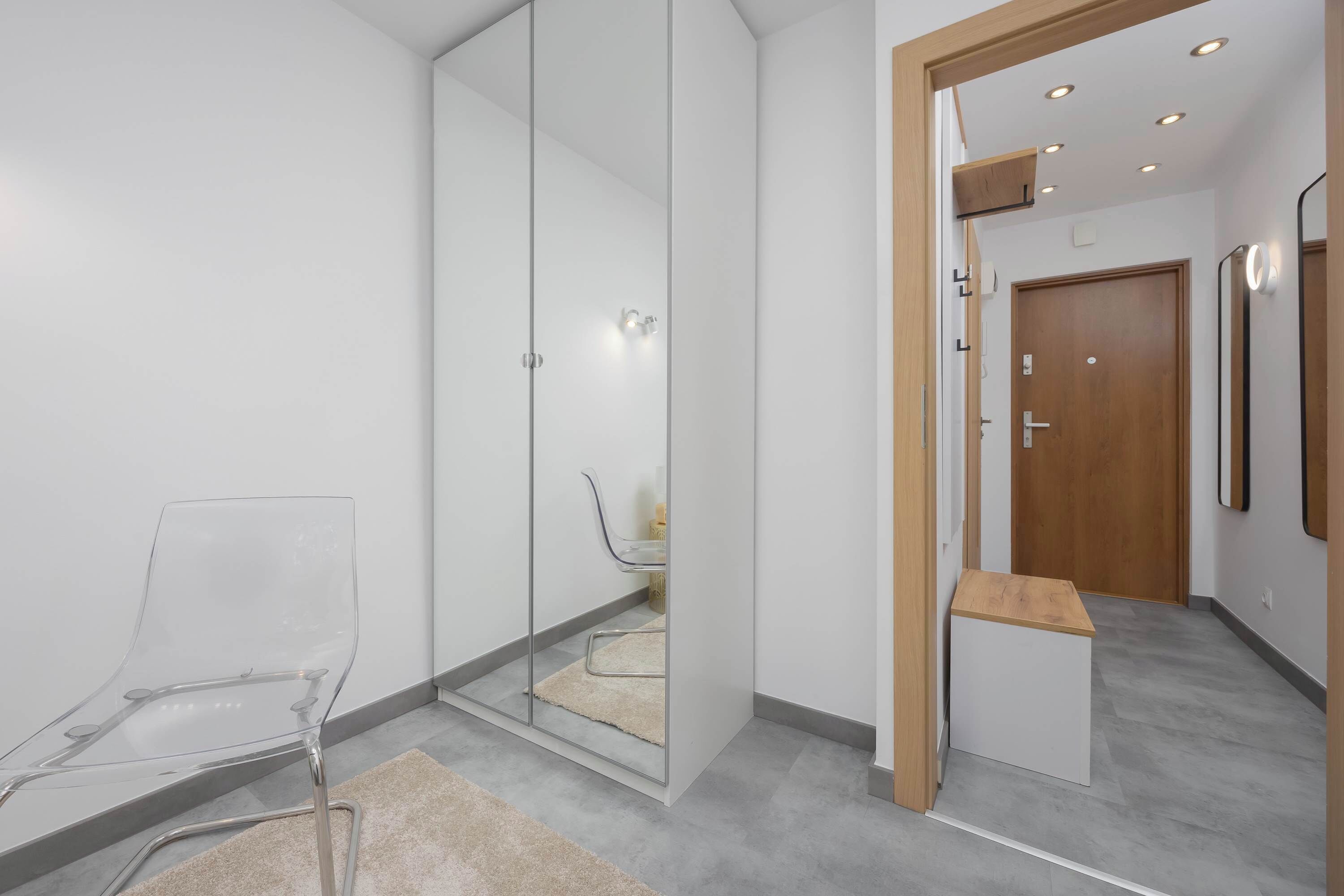 A modern hotel room with a glass partition, a clear chair, a small wooden table, and a grey carpeted floor.