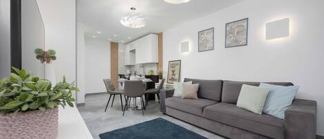 A modern living room with a grey sofa, a coffee table, and a dark rug. There is a dining area with chairs and a table in the background.
