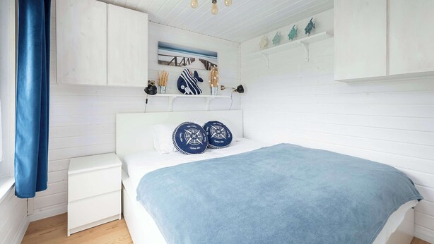 A bedroom with a bed, a nightstand, a headboard, a wall-mounted shelf with decorative items, and a ceiling light fixture.