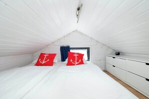 Ein Schlafzimmer mit schrägem Dach, einem Bett mit weißer Bettwäsche und roten Kissen mit Anker-Motiven, einer weißen Kommode und einem Fenster mit blauen Vorhängen.