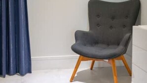 A room with a grey upholstered chair with wooden legs, a patterned blue rug, and a white wall. - Two Bedroom Cable Beach Condo With Patio (Nassau)