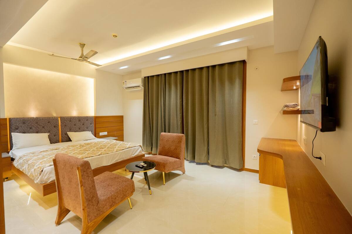 A modern hotel room with a bed, two armchairs, a small table, a ceiling fan, an air conditioning unit, a TV, and a large window with curtains.