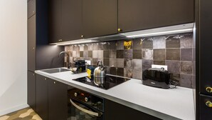 Cozinha moderna com armários escuros, bancada branca, chaleira de aço inoxidável, torradeira e forno embutido.