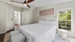 Un dormitorio con una cama grande, un ventilador de techo, una ventana con vista a follaje y una puerta que da a otra habitación.