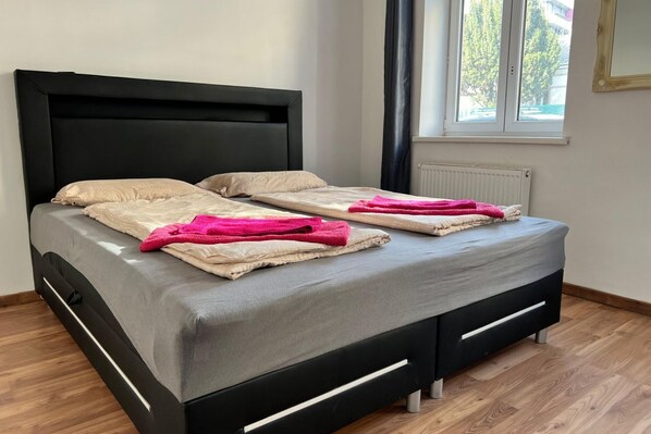 Una cama doble con cabecera negra, ropa de cama gris y dos toallas rosas.