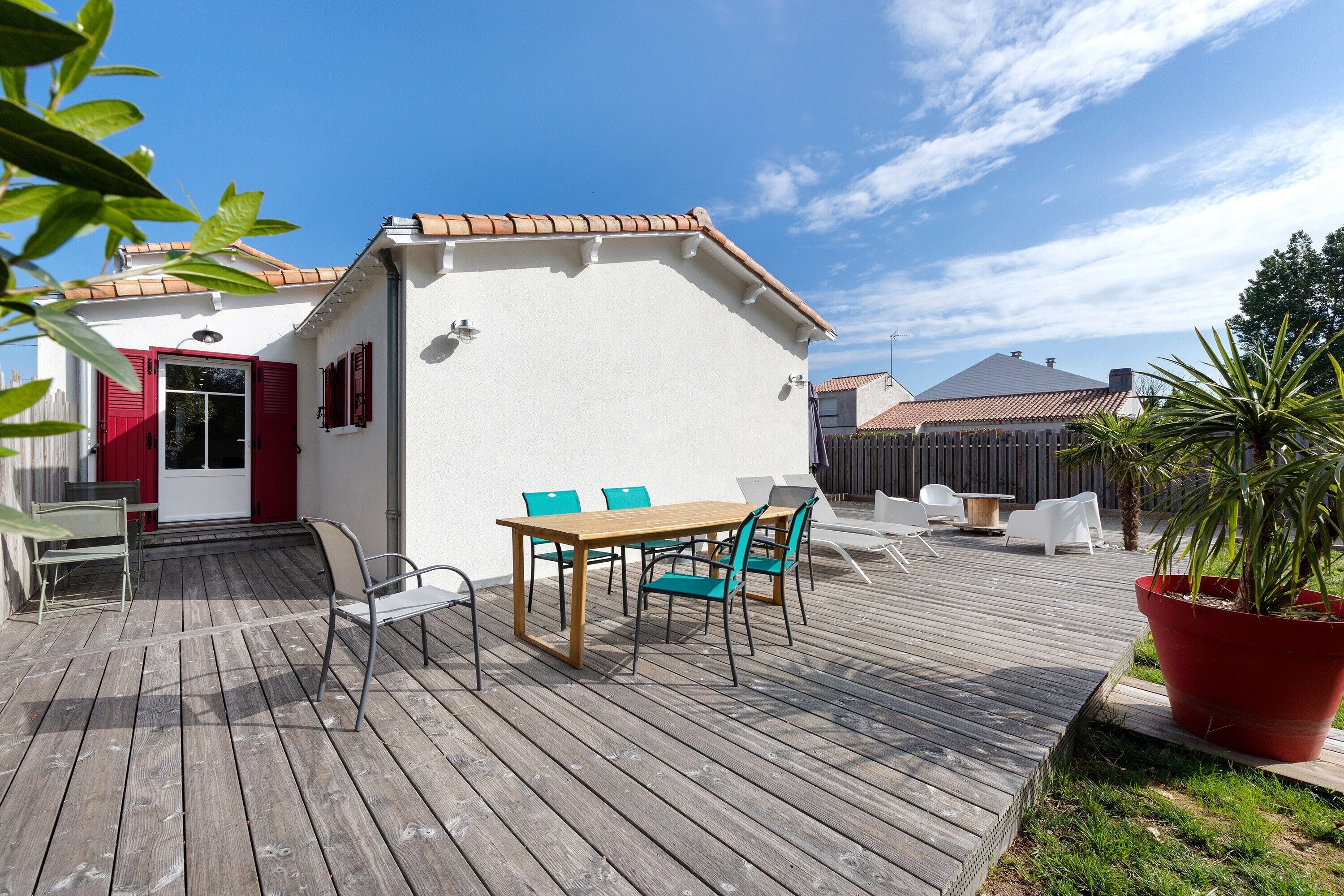 A wooden deck with a table and chairs, a potted plant, and a building with red shutters.
