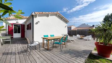 A wooden deck with a table and chairs, a potted plant, and a building with red shutters.