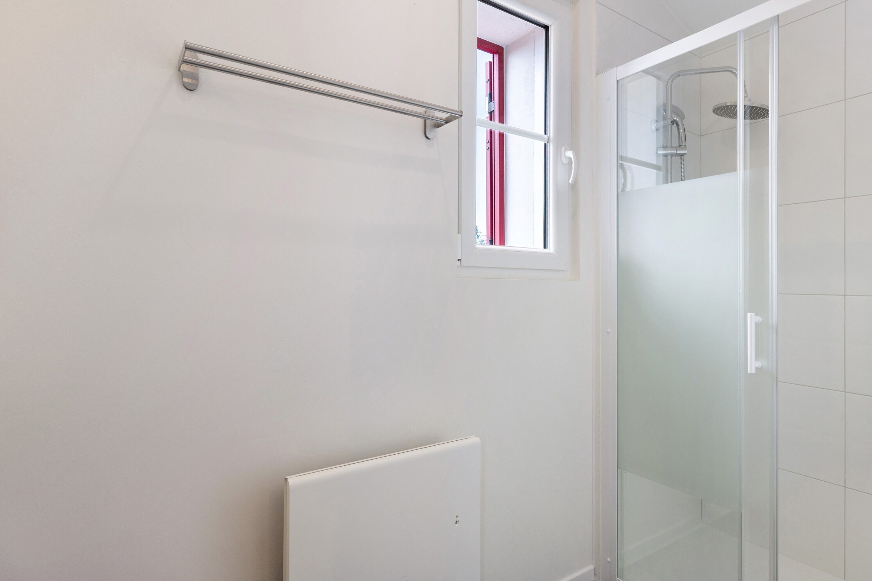 Un baño con cabina de ducha de vidrio, un radiador blanco y una ventana con marco rojo.