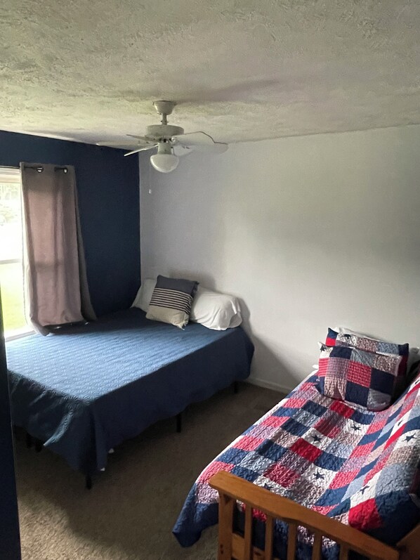 Un dormitorio con dos camas, una con colcha azul y la otra con cubrecama de colcha. Hay un ventilador de techo y una ventana con cortinas.