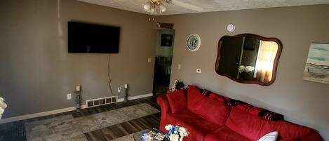 A living room with a red couch, a coffee table with magazines, a ceiling fan, a flat-screen TV, and a mirror on the wall.