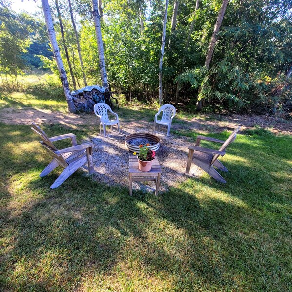 Un área de estar al aire libre con una fogata, dos sillas de plástico blancas y un banco de madera.