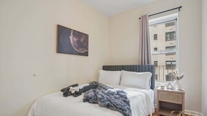 A bedroom with a bed, a bedside table, a window with curtains, and a painting on the wall.