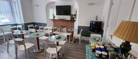 Un comedor con mesas redondas de cristal y sillas blancas, chimenea de ladrillo, televisor de pantalla plana y un reloj en la pared.