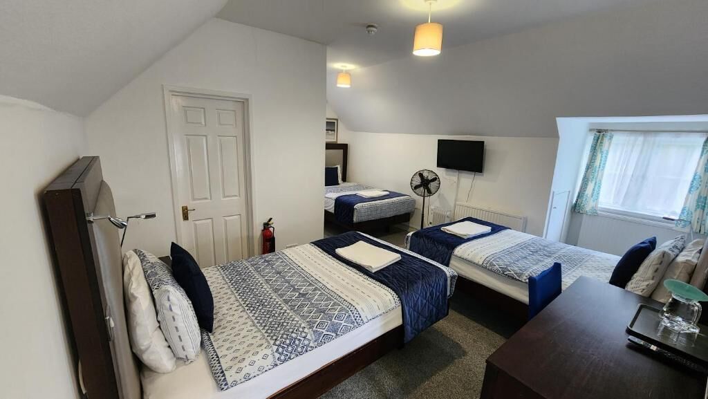 A room with two beds, a desk, a chair, a TV, a fan, and a window with curtains.