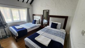 A room with two beds, a standing fan, and a window with curtains.