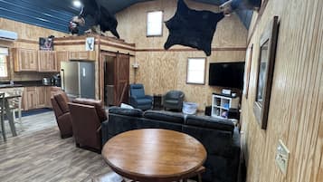 A rustic interior with a wooden dining table and chairs, a sofa, and a kitchen area with a refrigerator and cabinets.
