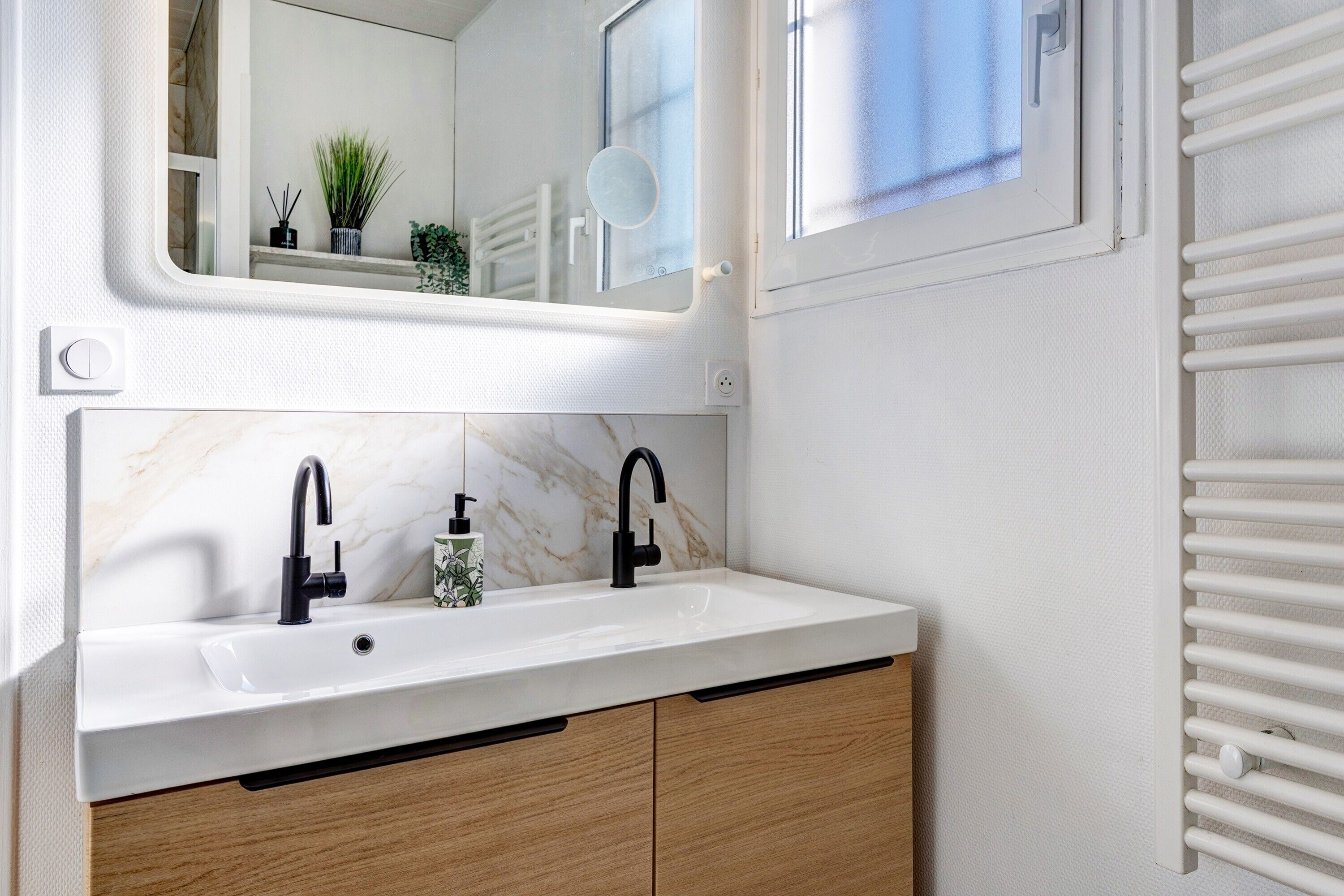 Ein Badezimmer mit weißem Waschbecken, schwarzen Wasserhähnen, Holzschränken, einem Spiegel und einem Fenster mit Jalousien.