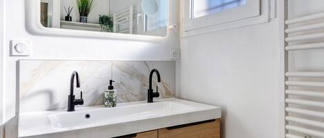 Ein Badezimmer mit weißem Waschbecken, schwarzen Wasserhähnen, Holzschränken, einem Spiegel und einem Fenster mit Jalousien.