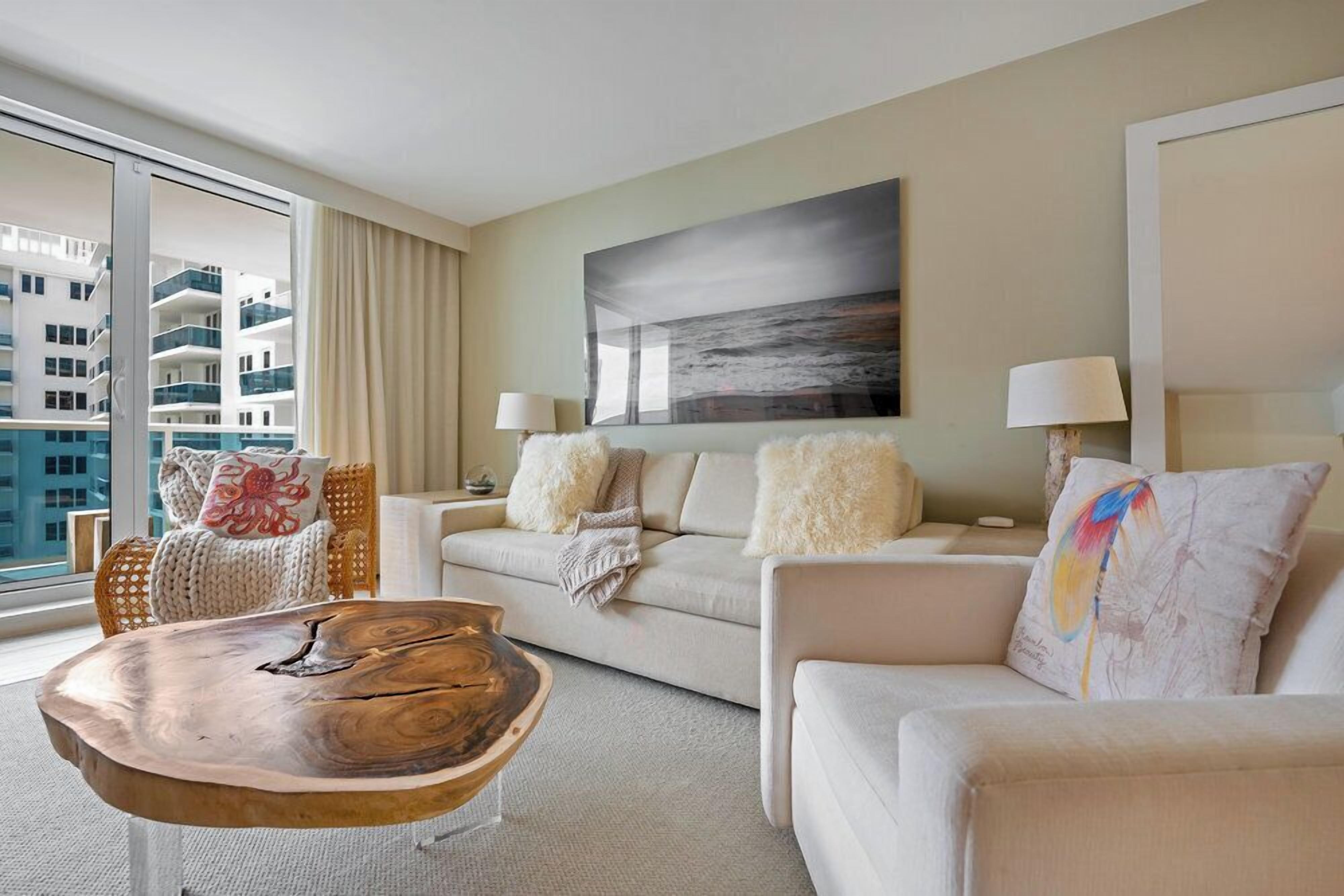 A modern hotel room with a large window, a white sofa, a wooden coffee table, and a painting on the wall.