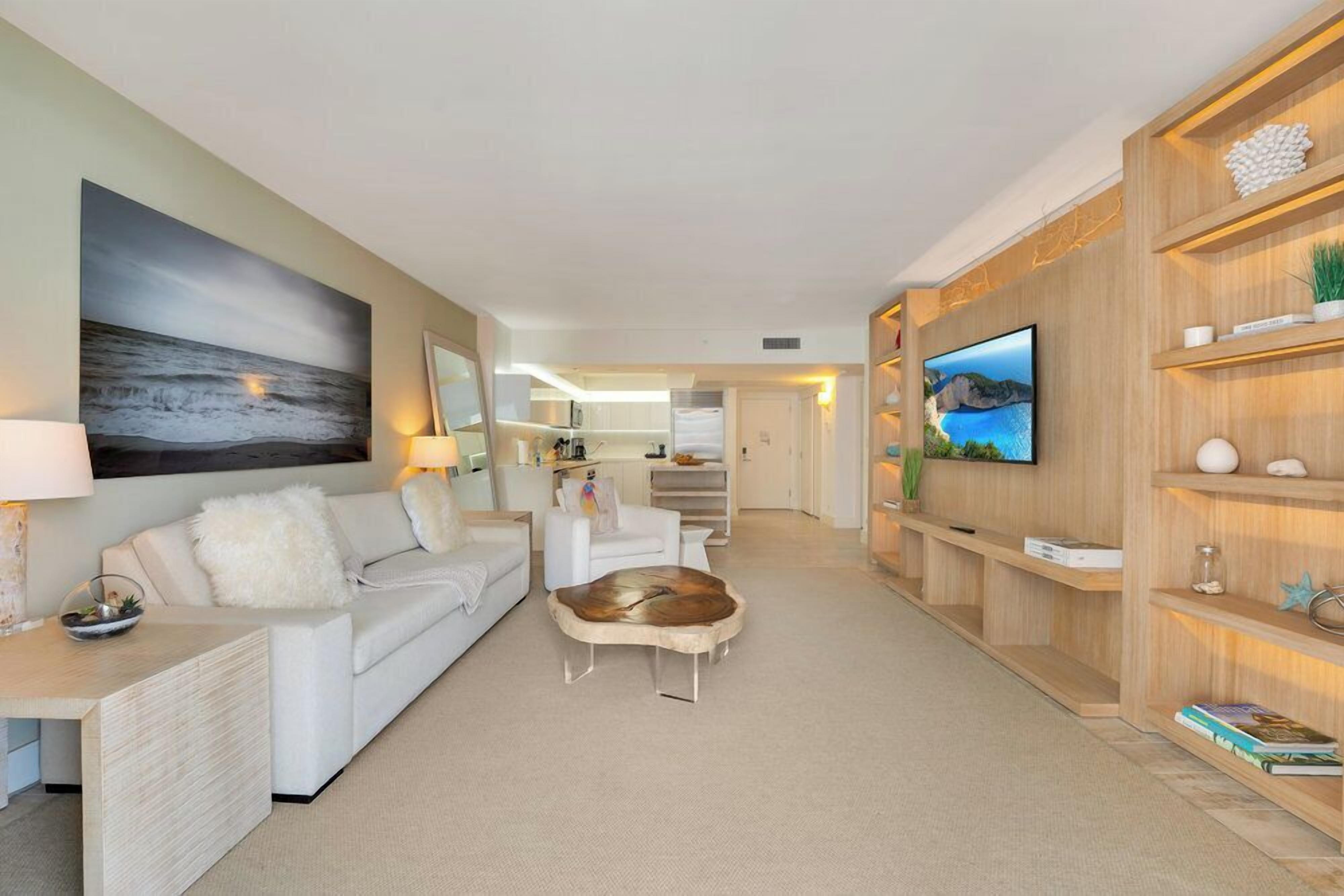 A modern living room with a sofa, coffee table, and TV. There is a kitchen in the background and a wooden shelf with decorative items.