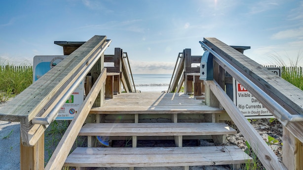 Una passerella in legno conduce alla spiaggia, con segnali di avvertimento ben visibili e una vista libera sull'oceano.