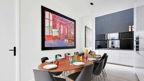 Un comedor moderno con una mesa de madera preparada para seis personas, rodeada de sillas negras. Un cuadro grande adorna la pared sobre la mesa, y se ve una cocina a la derecha.