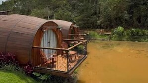 Eine zylindrische Holzhütte mit Balkon, gelegen an einem Gewässer, umgeben von üppiger Vegetation und Bäumen.