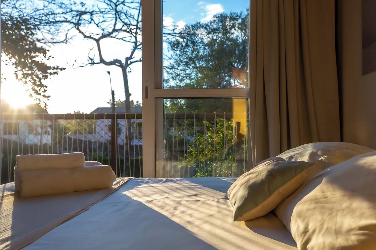 Une chambre avec une grande fenêtre, un lit avec des oreillers et une vue sur des arbres et une clôture.