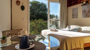 Ein Hotelzimmer mit einem Bett, einem Tisch mit einer Kaffeetasse, einem Stuhl und einem Balkon mit Blick auf Bäume.