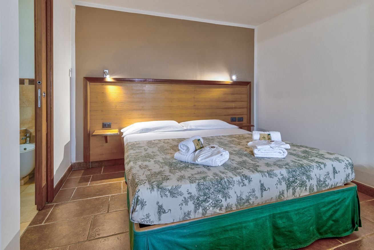 Habitación de hotel con cama, cabecera de madera, falda verde y baño visible a través de una puerta abierta.