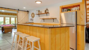 A kitchen with wooden cabinets, a built-in island with stools, a refrigerator, and a pool table in the background. - Ski, Hike & Game: White Mountains Family Retreat (Campton)