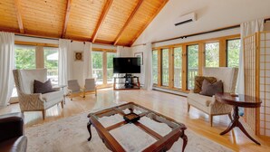 A spacious living room with wooden floors, large windows, a television, and comfortable seating. - Ski, Hike & Game: White Mountains Family Retreat (Campton)