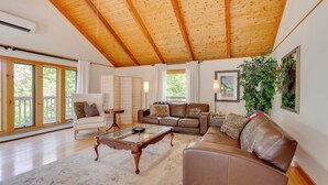 A spacious living room with a high wooden ceiling, large windows, a brown leather sofa, a white armchair, and a glass coffee table. - Ski, Hike & Game: White Mountains Family Retreat (Campton)