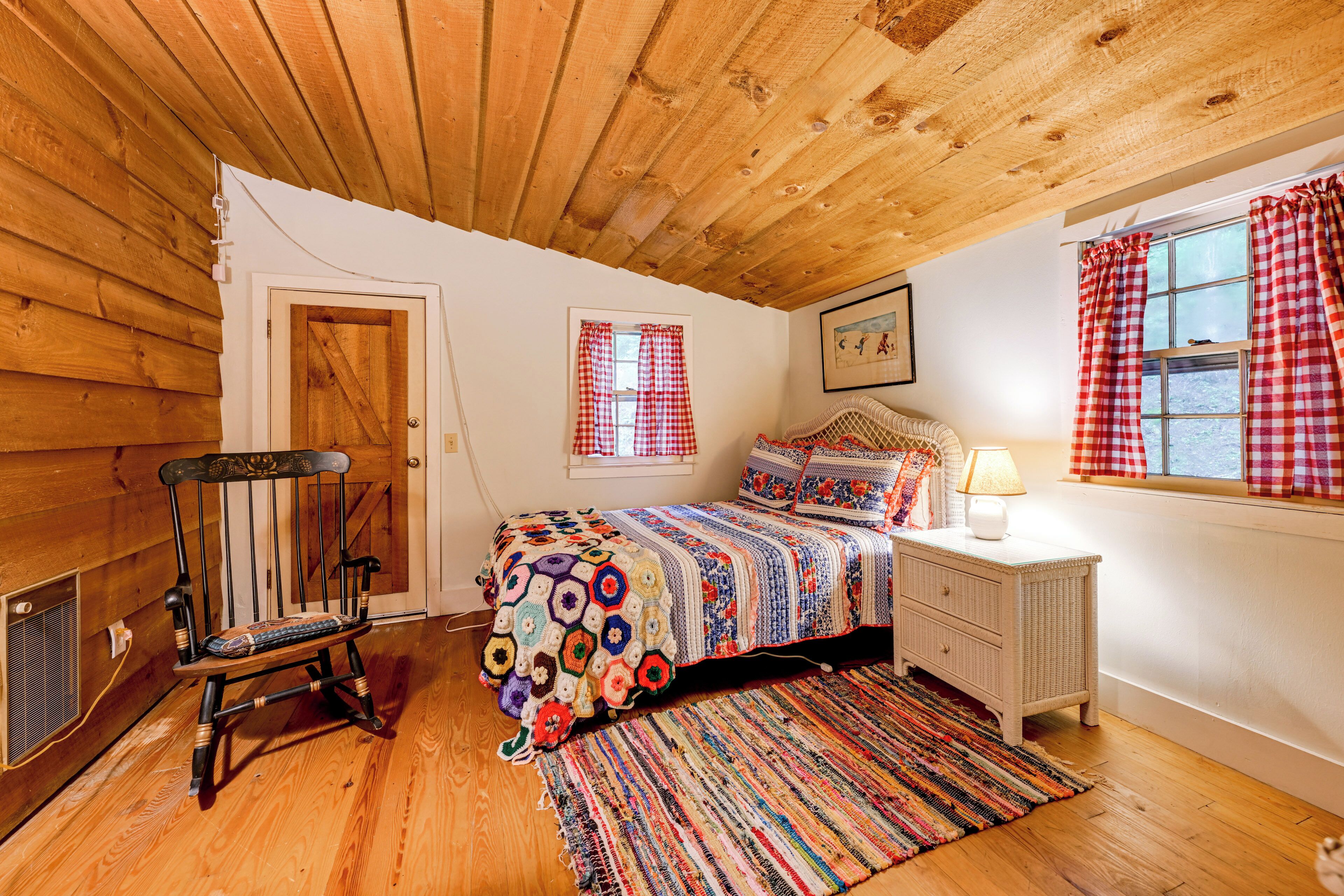 Un dormitorio acogedor con techo de madera, una cama con colcha colorida, un banco y una ventana con cortinas a cuadros.