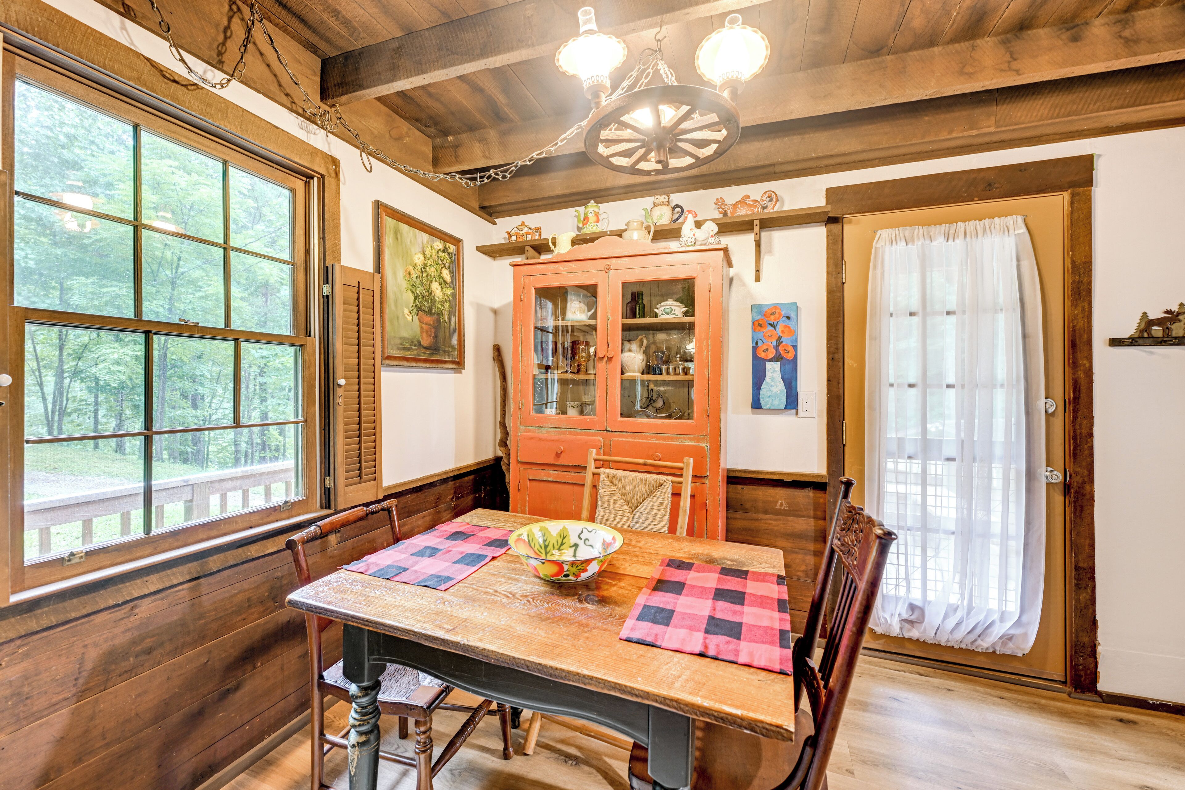 Una mesa de comedor de madera con un bol y manteles individuales a cuadros, rodeada de sillas, en una habitación con paredes de madera y un ventanal grande.