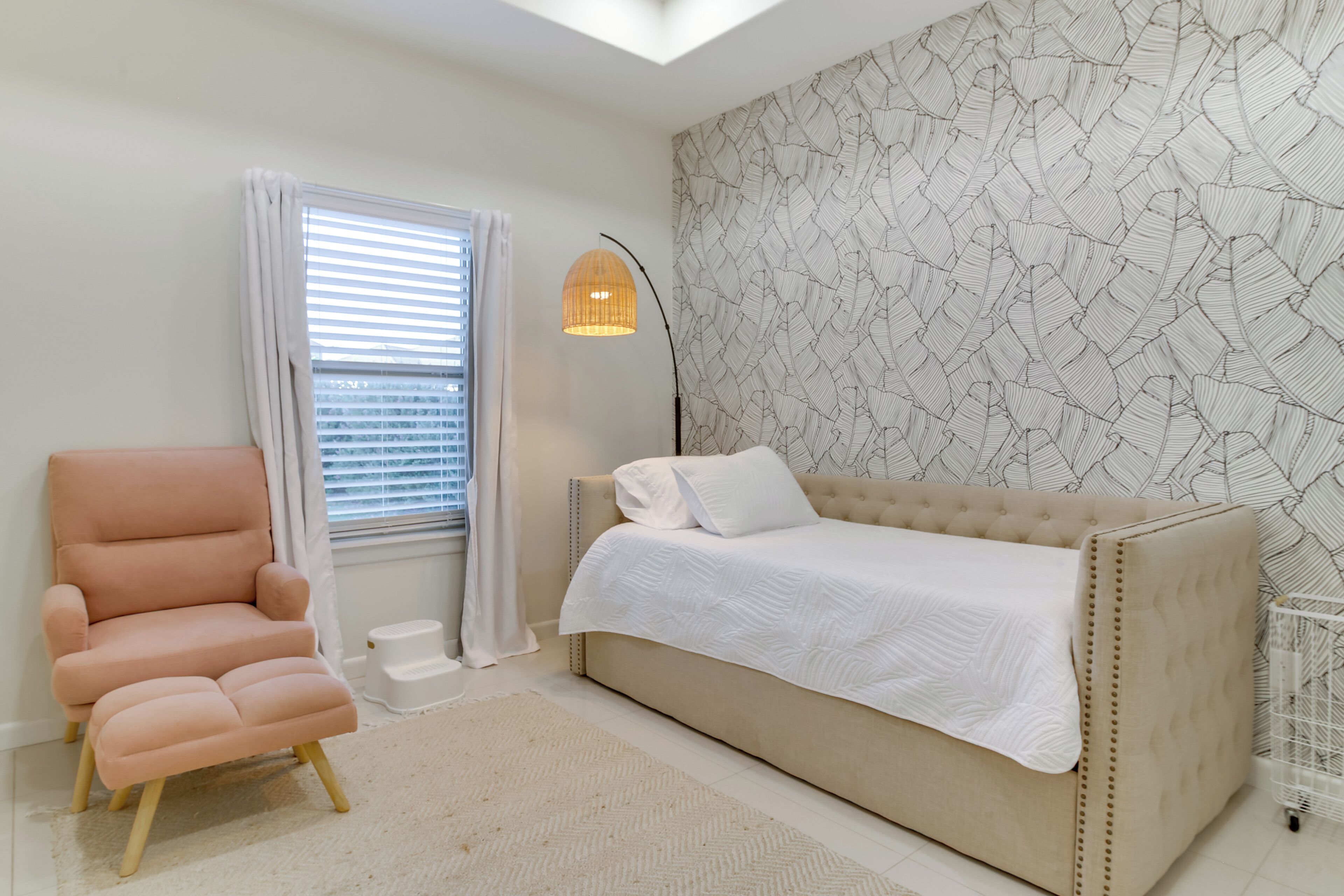 Une chambre avec un canapé beige, un fauteuil rose, un lit blanc, un mur à motifs, une lampe sur pied et une fenêtre avec des stores.
