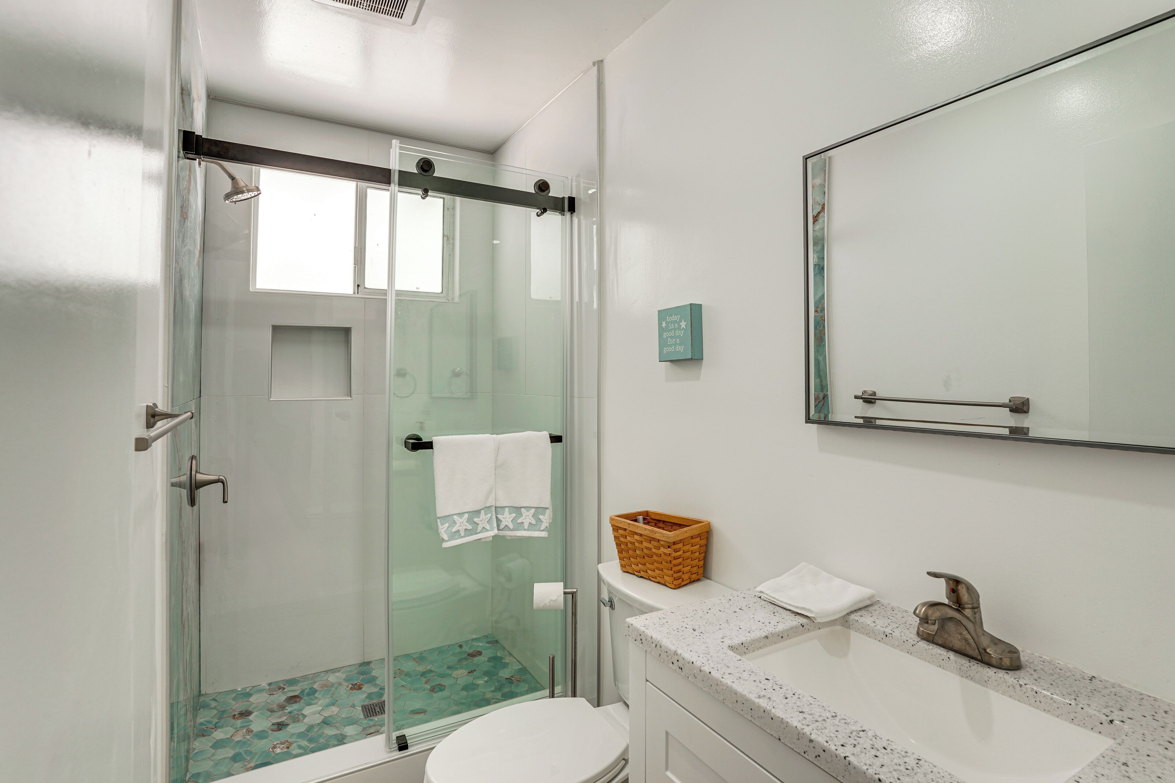 A bathroom with a glass shower enclosure, a white toilet, a sink with a speckled countertop, and a mirror above the sink.