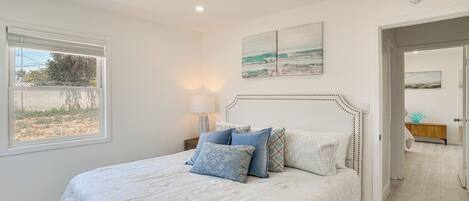 A bedroom with a large bed, a window, a bedside table with a lamp, and a door leading to another room.
