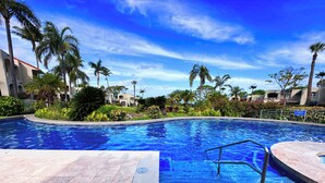 Una piscina con cielo azul despejado, rodeada de palmeras y un jardín bien cuidado.