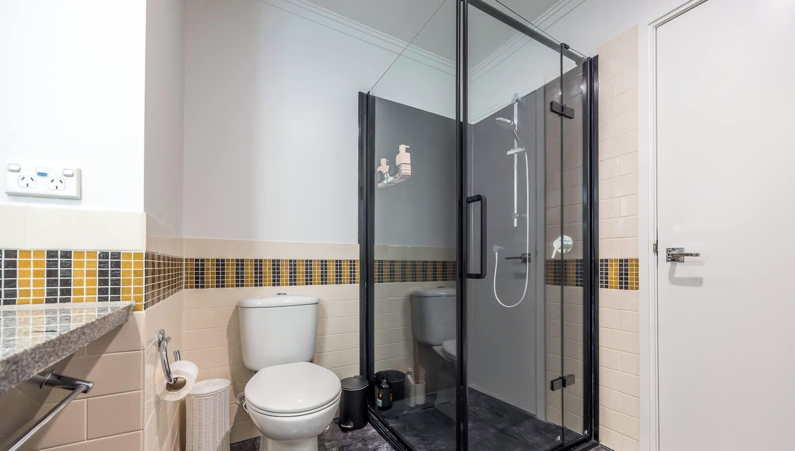 Une salle de bain comprenant des toilettes, une douche fermée par une paroi de verre et un mur recouvert de carreaux.