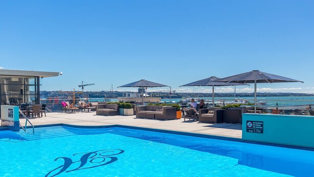 A rooftop pool with lounge chairs and umbrellas, a clear view of the sea, and a sign indicating no running or jumping. - Relaxing 1BR CBD Retreat with Rooftop pool & Gym (Auckland)