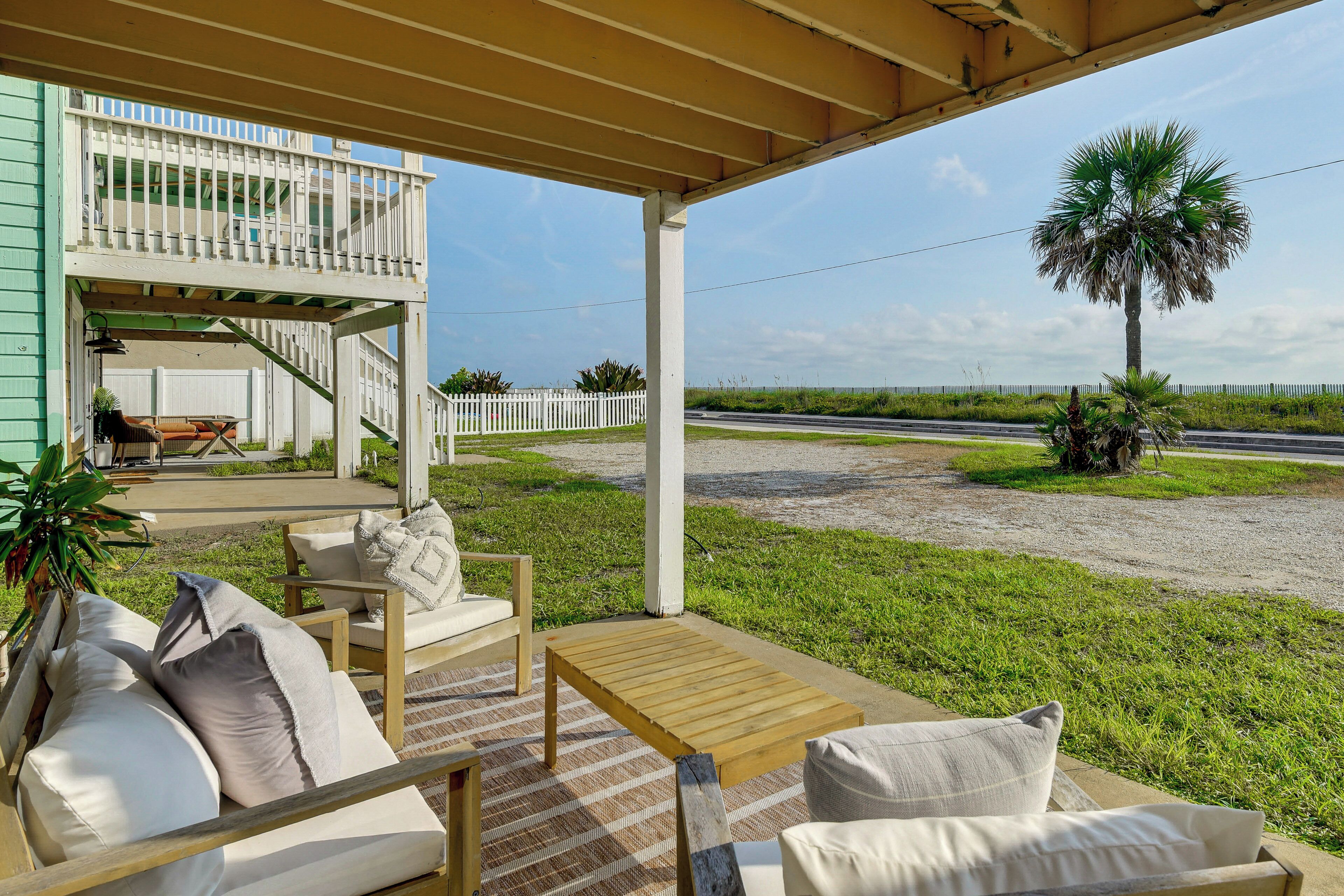A covered outdoor seating area with a wooden table and chairs, a staircase to an upper level, and a view of the road and palm trees.
