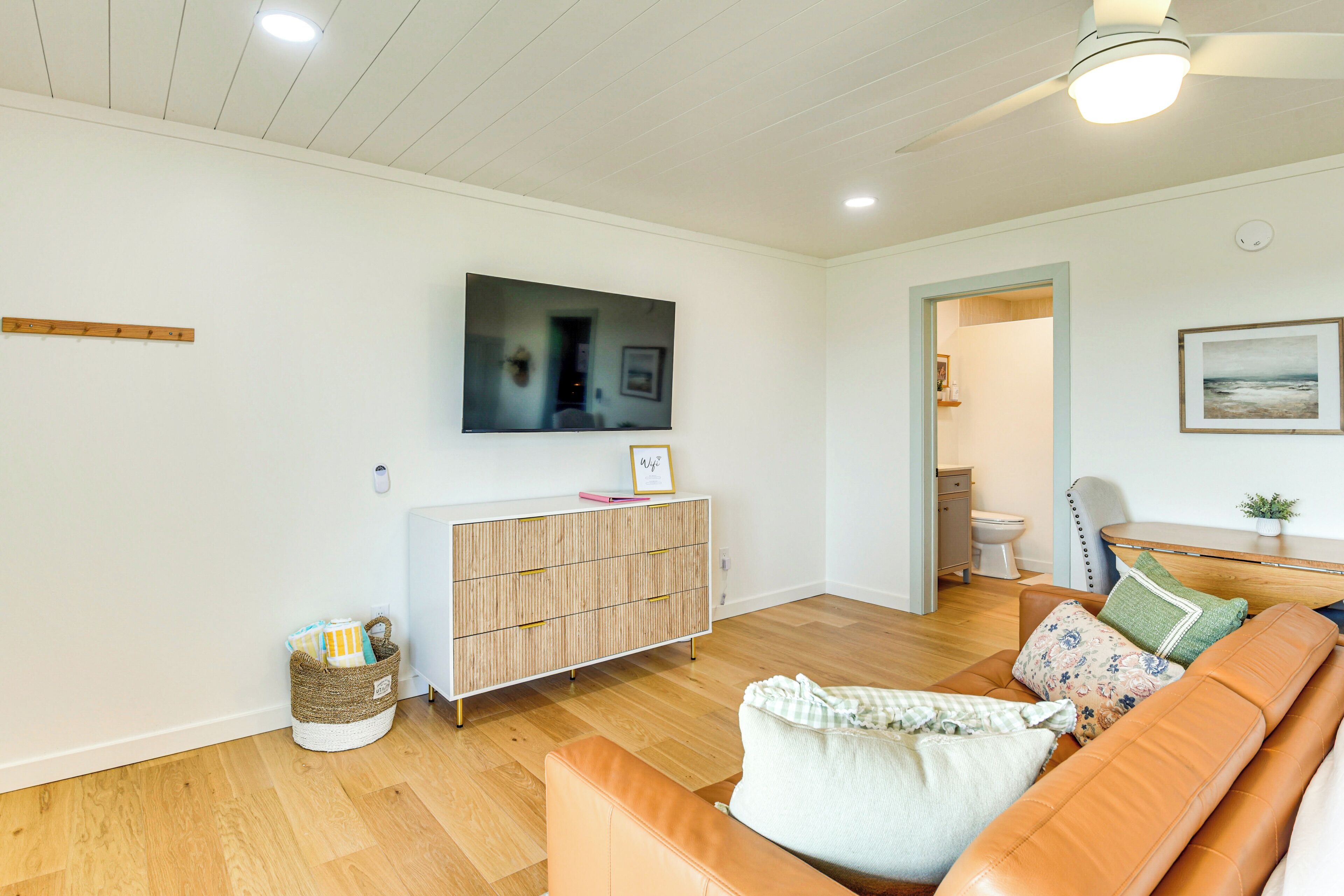 Un salon avec un plancher en bois, un canapé, un téléviseur fixé au mur et un meuble avec des tiroirs.