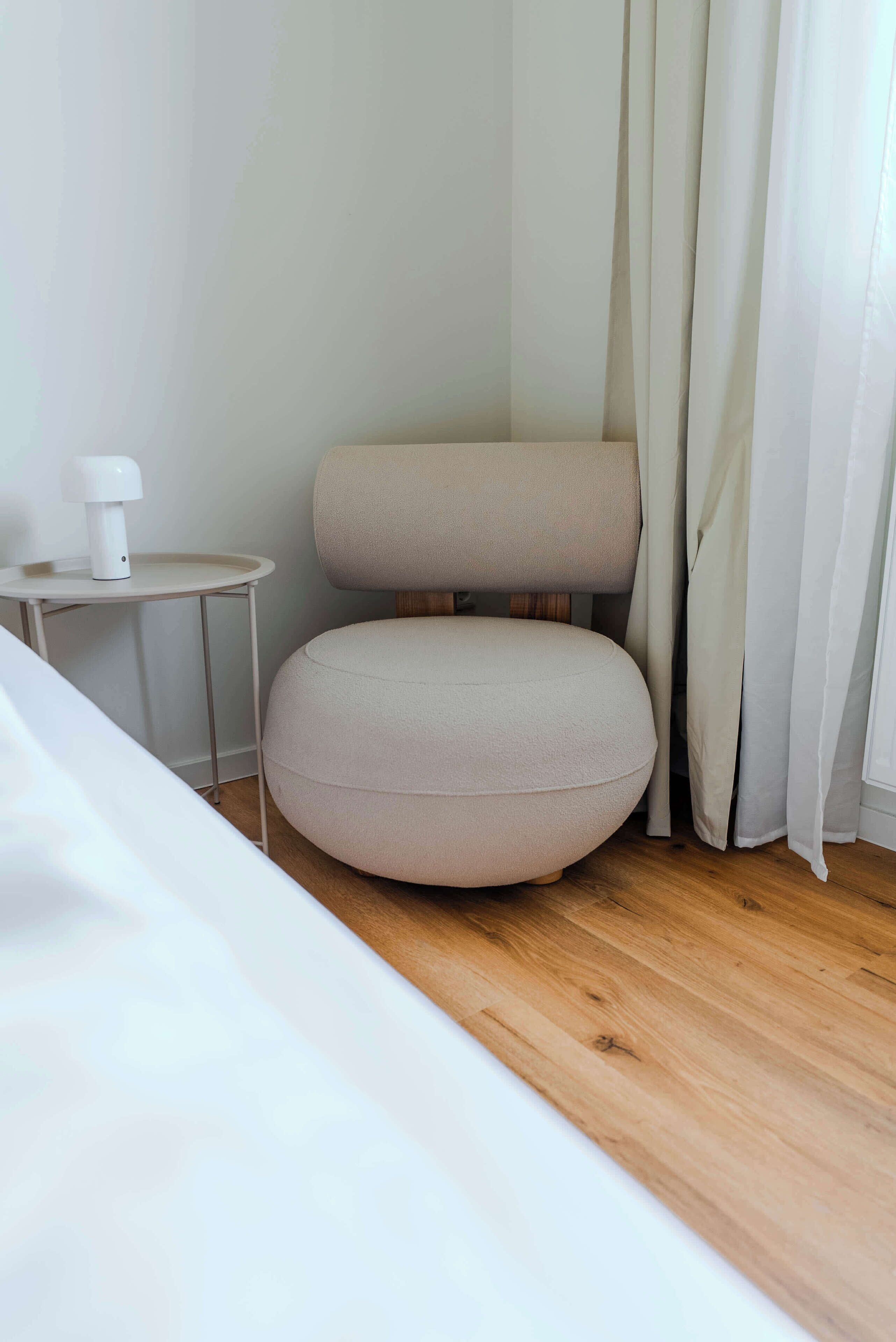 A modern room with a round, light-colored ottoman, a small side table with a white lamp, and a wooden floor.