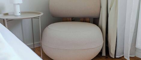 A modern room with a round, light-colored ottoman, a small side table with a white lamp, and a wooden floor.